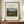 May include: Framed art print of Henri Rousseau's pastoral landscape painting featuring four young girls in red dresses walking through tall grass, with industrial buildings and smokestacks in the background under a cloudy sky. The cream-matted print in a thin brown frame sits on a wooden sideboard next to a small white ceramic vase and stacked books against white wainscoting.