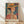 May include: Framed art print leaning against white wall on light wood floor. Colorful portrait shows woman wearing orange wide-brimmed hat with dark band, geometric patterned clothing in warm oranges and blues. Background features vibrant tropical and geometric patterns in teal, orange, and green. Artist name printed at bottom in dark text on cream background. Natural wood frame, soft lighting creates diagonal shadows on white fabric backdrop.