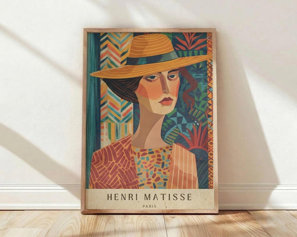 May include: Framed art print leaning against white wall on light wood floor. Colorful portrait shows woman wearing orange wide-brimmed hat with dark band, geometric patterned clothing in warm oranges and blues. Background features vibrant tropical and geometric patterns in teal, orange, and green. Artist name printed at bottom in dark text on cream background. Natural wood frame, soft lighting creates diagonal shadows on white fabric backdrop.