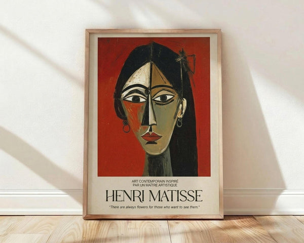 May include: Framed poster print featuring cubist-style portrait of woman with long black hair against red background, displayed on light wooden floor against white wall with natural window light casting diagonal shadows. Artwork shows geometric facial features in earth tones with text crediting Henri Matisse below the image in cream-colored matting within natural wood frame.