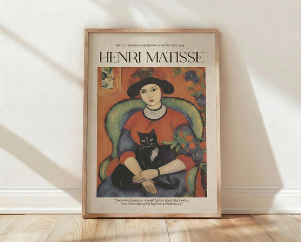 May include: Framed Henri Matisse art print leaning against white wall on light wooden floor. Poster features woman in red top and black hat sitting in green chair, holding black and white cat. Warm orange background with floral elements. Natural wood frame, cream matting, inspirational quote below image. Soft natural lighting creates gentle shadows on textured white wall.