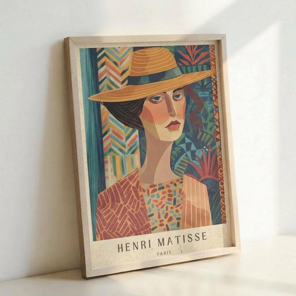 A colorful art print in a light wooden frame showing a stylized portrait of a woman wearing a wide-brimmed yellow hat. The artwork features geometric patterns and warm tones including orange, teal, and coral, with decorative elements surrounding the figure. Text reading "HENRI MATISSE PARIS" appears at the bottom. The framed print leans against a white wall.