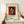 May include: Hands positioning a framed Matisse-style portrait print featuring a woman's face in cubist style with geometric shapes, red background, and black hair. The wooden frame sits on a light wood surface with a white wrapped gift box nearby. The poster shows French text and an inspirational quote below the colorful modernist artwork.