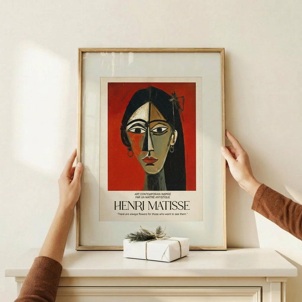 May include: Hands positioning a framed Matisse-style portrait print featuring a woman's face in cubist style with geometric shapes, red background, and black hair. The wooden frame sits on a light wood surface with a white wrapped gift box nearby. The poster shows French text and an inspirational quote below the colorful modernist artwork.