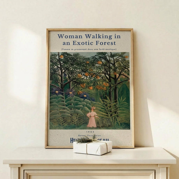 May include: Framed art print displaying a lush jungle scene with dense green foliage, tropical plants, and a small figure in pink walking through an exotic forest. The vintage-style poster features rich emerald and forest green tones with orange accents, displayed in a thin wooden frame on a light wood surface alongside a small white wrapped gift with natural twine and evergreen sprig.