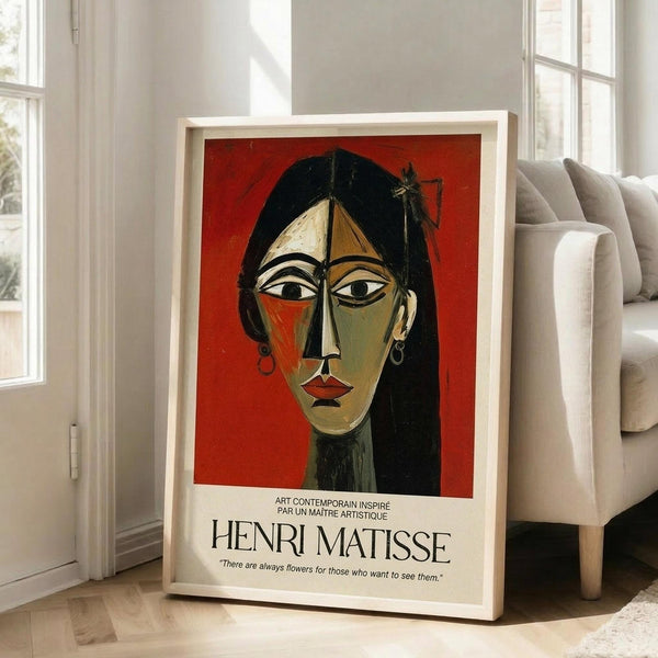 May include: Framed poster print featuring an abstract portrait of a woman with long black hair, geometric facial features in green and orange tones against a red background, attributed to Henri Matisse with French text and quote about flowers below the artwork, displayed on hardwood floor next to beige upholstered chair in bright room with white walls and windows