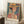 May include: A framed art poster featuring a woman wearing an orange wide-brimmed hat against a vibrant background of geometric patterns in blues, greens, and warm tones. The artwork shows bold colors and stylized forms with "HENRI MATISSE PARIS" text at bottom. The poster leans against a white wall corner next to a wooden side table with coffee cup, on a textured white rug with neutral bedding visible.