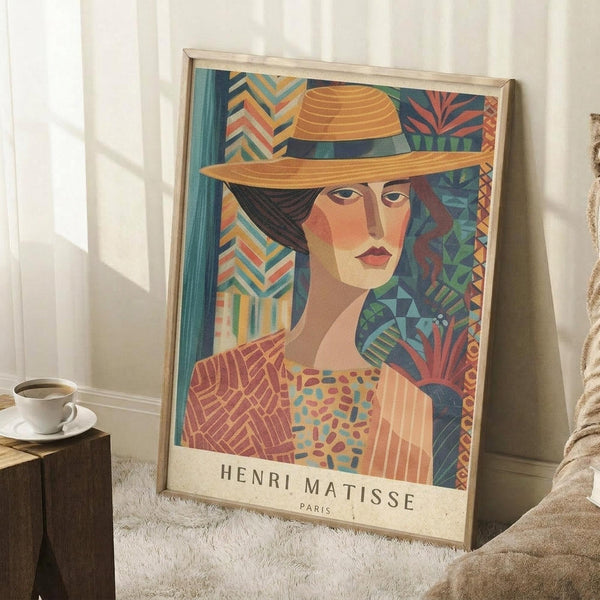 May include: A framed art poster featuring a woman wearing an orange wide-brimmed hat against a vibrant background of geometric patterns in blues, greens, and warm tones. The artwork shows bold colors and stylized forms with "HENRI MATISSE PARIS" text at bottom. The poster leans against a white wall corner next to a wooden side table with coffee cup, on a textured white rug with neutral bedding visible.