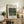 May include: A framed art print displaying Henri Rousseau's jungle painting featuring a woman in pink dress walking through lush green tropical foliage. The poster has white matting with exhibition-style text. The wooden-framed print leans against a white wall on a rustic wood sideboard, styled with a fern plant, books, ceramic vase, and cream upholstered chair visible in the cozy living room setting.