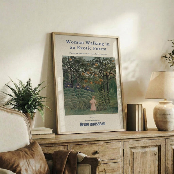 May include: A framed art print displaying Henri Rousseau's jungle painting featuring a woman in pink dress walking through lush green tropical foliage. The poster has white matting with exhibition-style text. The wooden-framed print leans against a white wall on a rustic wood sideboard, styled with a fern plant, books, ceramic vase, and cream upholstered chair visible in the cozy living room setting.