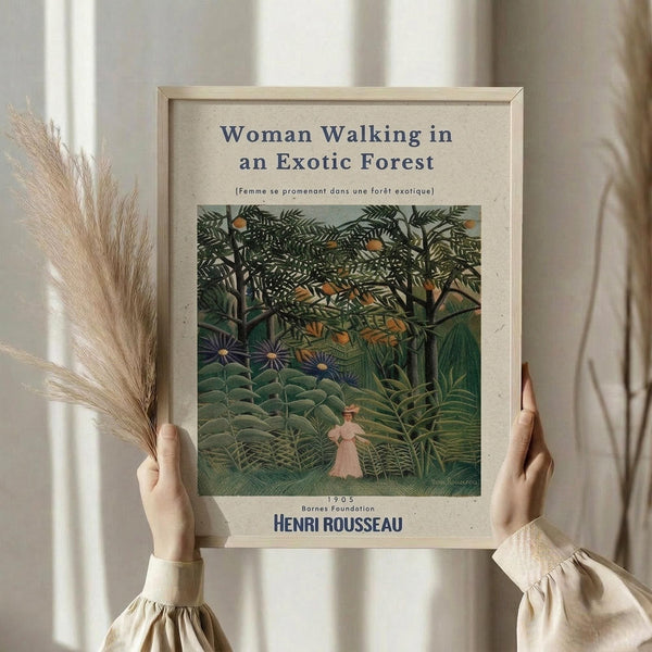 May include: Hands holding a framed art print of Henri Rousseau's jungle painting showing a woman in pink dress walking through lush green tropical forest with dense foliage and exotic plants. The print has museum-style text layout with title and artist name. Person wears cream colored ruffled sleeves and holds dried wheat stems, standing against white curtained window background in soft natural lighting.