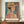 May include: Framed poster print featuring a woman wearing a wide-brimmed orange hat in colorful geometric Art Deco style with botanical patterns in the background, displayed on a wooden sideboard next to a white ceramic vase and stack of books against white wainscoting wall. The artwork shows warm earth tones, blues, and coral colors with "HENRI MATISSE PARIS" text at bottom.