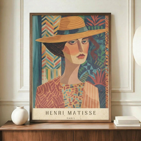 May include: Framed poster print featuring a woman wearing a wide-brimmed orange hat in colorful geometric Art Deco style with botanical patterns in the background, displayed on a wooden sideboard next to a white ceramic vase and stack of books against white wainscoting wall. The artwork shows warm earth tones, blues, and coral colors with "HENRI MATISSE PARIS" text at bottom.