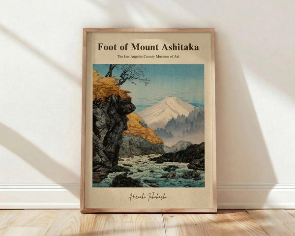 May include: Framed vintage Japanese woodblock print showing Mount Fuji in the distance with autumn foliage, rocky cliffs, and a rushing stream in the foreground. The artwork features muted blues, yellows, and grays in traditional ukiyo-e style. Light wood frame sits against white wall on blonde hardwood floor with soft natural lighting creating shadows.