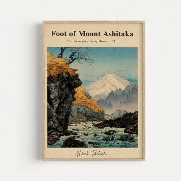 Framed vintage Japanese woodblock print showing Mount Fuji in the distance with snow-capped peak against blue sky. In foreground, a rushing stream flows over dark rocks between autumn trees with golden foliage. Rocky cliffs and misty mountains create layers of depth. Natural wood frame against white wall. Art attributed to Hiroaki Takahashi with Los Angeles County Museum of Art credit.