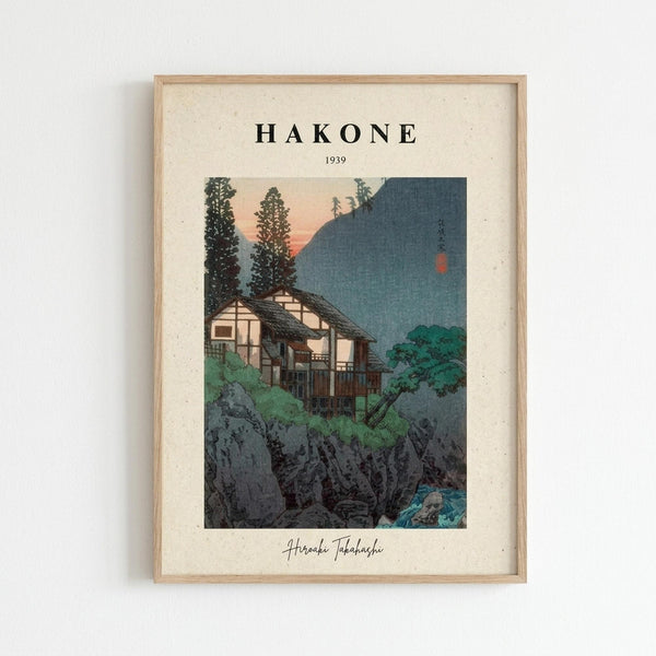 May include: Framed vintage Japanese woodblock print poster mounted on white wall. Shows traditional mountain village scene with wooden houses nestled among tall evergreen trees and rocky cliffs. Features muted blue-green mountains, peachy sunset sky, and small red moon. Text reads "HAKONE 1939" at top with artist signature below. Natural wood frame with cream matting against textured off-white wall background.