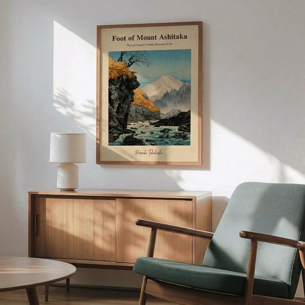 May include: Japanese woodblock print poster showing Mount Fuji landscape with autumn trees, rocky stream, and misty mountains in blues, oranges and earth tones, displayed in wooden frame above mid-century modern credenza with white ceramic lamp, next to sage green upholstered armchair with wooden arms in sunlit room with white walls.