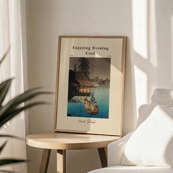 May include: Framed vintage Japanese woodblock print showing people in a boat by a waterside pavilion at dusk, displayed on round wooden side table next to beige upholstered chair. Print features blue water, traditional architecture, and evening sky in muted tones. Natural lighting creates soft shadows on cream wall with plant leaves visible in corner.