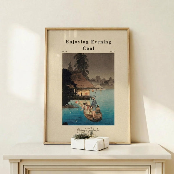 May include: Framed Japanese woodblock print showing people in a boat near a traditional house by water at evening, with mountains in background. Print has beige matting in light wood frame, displayed on white wooden surface next to small wrapped gift with twine and greenery. Soft natural lighting creates shadows on cream wall behind.