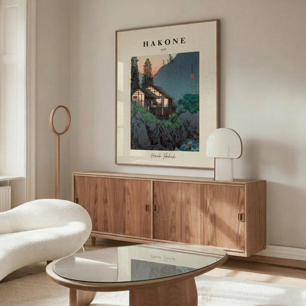 May include: Framed vintage Japanese woodblock print poster showing traditional houses nestled among green trees and rocky landscape at dusk, displayed above a mid-century modern walnut credenza. Scene includes cream upholstered chair, glass-top coffee table, and modern table lamp in bright, minimalist living room with white walls.