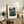 May include: Framed vintage-style Japanese woodblock print showing traditional houses nestled among trees with mountain backdrop in muted blues and greens, displayed on rustic wooden sideboard alongside fern plant, ceramic vase, and stacked books in warm neutral living room setting.