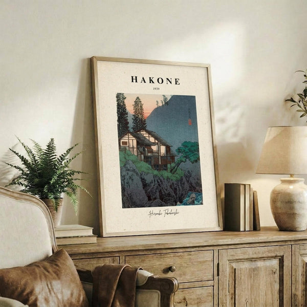 May include: Framed vintage-style Japanese woodblock print showing traditional houses nestled among trees with mountain backdrop in muted blues and greens, displayed on rustic wooden sideboard alongside fern plant, ceramic vase, and stacked books in warm neutral living room setting.
