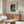 May include: Japanese woodblock-style poster print hanging above wooden sideboard in modern living room. Artwork shows lakeside scene with traditional house, boats, and evening sky in blues and earth tones. Room features cream chair, glass coffee table, white table lamp, and wooden floor lamp. Neutral walls and natural lighting create serene atmosphere.