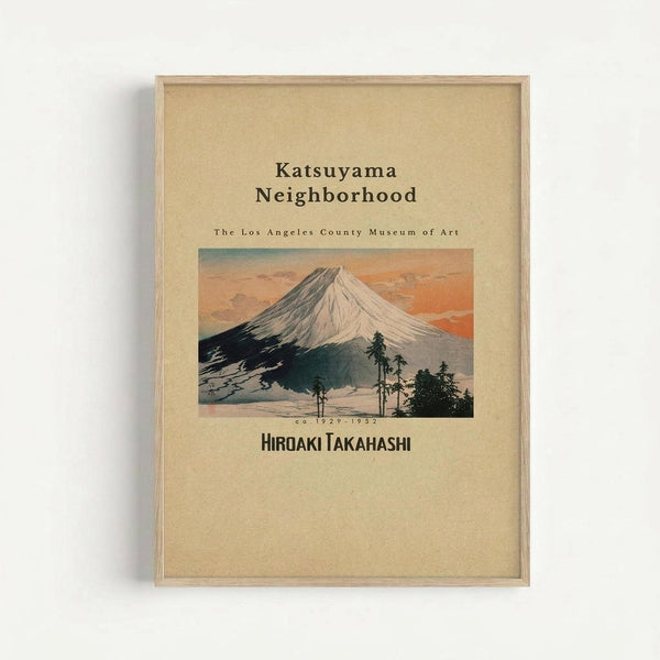 Framed vintage Japanese woodblock print featuring Mount Fuji with snow-capped peak against orange and blue sunset sky. Dark silhouettes of pine trees in foreground, with text reading "Katsuyama Neighborhood" and "The Los Angeles County Museum of Art" above the mountain scene. Artist name "Hiroaki Takahashi" appears below with dates "ca. 1929-1932". Print has cream-colored background with natural wood frame, displayed against white wall.
