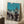 May include: Framed vintage Japanese woodblock print showing silhouetted figures dancing with lanterns against a turquoise and cream gradient sky. The artwork depicts traditional Obon festival dancers in flowing robes carrying illuminated paper lanterns. Print is displayed in a light wood frame, leaning against white paneled walls on a textured cream rug, with a coffee cup on a wooden block nearby and soft natural lighting creating gentle shadows.