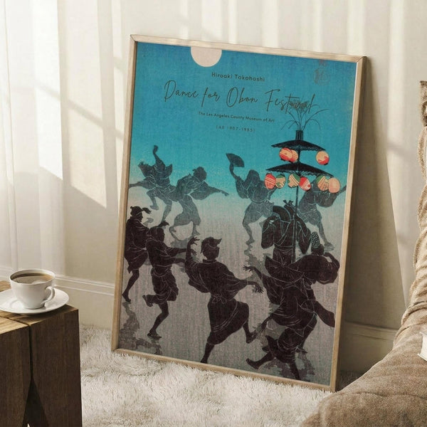May include: Framed vintage Japanese woodblock print showing silhouetted figures dancing with lanterns against a turquoise and cream gradient sky. The artwork depicts traditional Obon festival dancers in flowing robes carrying illuminated paper lanterns. Print is displayed in a light wood frame, leaning against white paneled walls on a textured cream rug, with a coffee cup on a wooden block nearby and soft natural lighting creating gentle shadows.