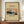 May include: Framed vintage Japanese woodblock print showing Mount Fuji with snow-capped peak against orange sunset sky, dark foreground with pine trees. Museum exhibition poster with cream background, brown wooden frame displayed on walnut credenza beside white ceramic vase and stacked books against white paneled wall.