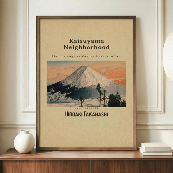 May include: Framed vintage Japanese woodblock print showing Mount Fuji with snow-capped peak against orange sunset sky, dark foreground with pine trees. Museum exhibition poster with cream background, brown wooden frame displayed on walnut credenza beside white ceramic vase and stacked books against white paneled wall.