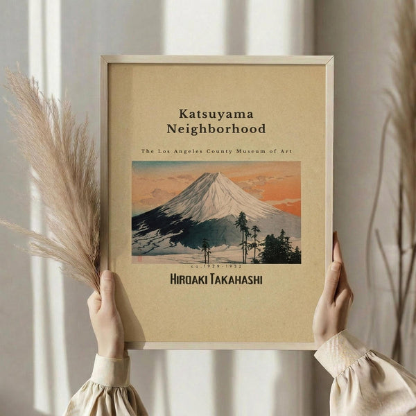 May include: Hands holding a framed vintage Japanese woodblock print featuring Mount Fuji with orange sunset sky and dark foreground trees. The beige poster has museum exhibition text and artist name Hiroaki Takahashi. Natural wood frame against soft neutral background with dried pampas grass visible on left side.