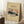 May include: Framed vintage-style poster featuring Mount Fuji woodblock print in earth tones, leaning against white wall corner beside wooden side table with coffee cup, on textured cream rug next to beige blanket