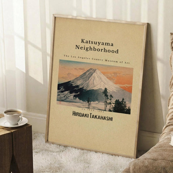 May include: Framed vintage-style poster featuring Mount Fuji woodblock print in earth tones, leaning against white wall corner beside wooden side table with coffee cup, on textured cream rug next to beige blanket