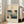 May include: Framed Japanese woodblock print poster showing sailing boats with white sails on blue water, displayed on light wooden floor against white wall corner next to beige upholstered chair. Natural sunlight streams through tall windows creating soft shadows. The artwork features traditional ukiyo-e style with cream matting in light wood frame.