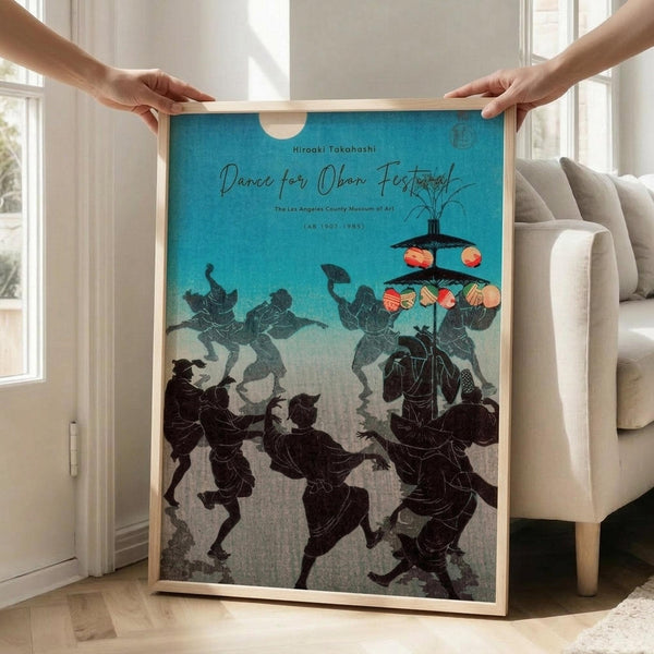 May include: Hands holding a framed poster with turquoise background showing silhouetted figures dancing around lanterns in traditional Japanese style. The artwork depicts dark figures in motion with colorful paper lanterns above. Light wooden frame against bright living room with beige sofa and large windows creating natural lighting on wooden floors.