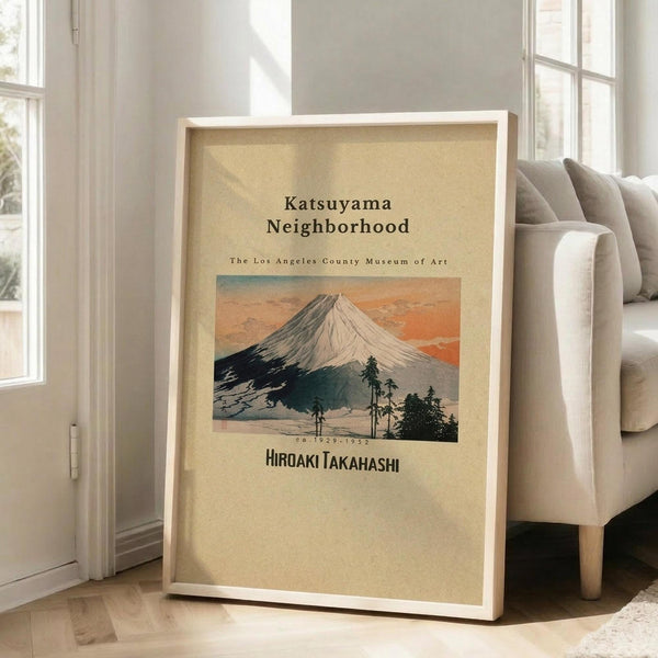 May include: Framed vintage-style poster featuring Mount Fuji with snow-capped peak against orange sunset sky, silhouetted trees in foreground, cream background with black text reading "Katsuyama Neighborhood" and "The Los Angeles County Museum of Art" and artist name, displayed in light wood frame leaning against white wall next to beige upholstered chair in bright living room with large windows.