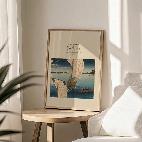 May include: Framed vintage Japanese woodblock print showing boats with white sails on blue water under evening sky with full moon. Light wood frame displayed on round wooden side table next to beige upholstered chair. Soft natural lighting creates gentle shadows on cream wall. Green plant leaves visible in corner of minimalist room setting.