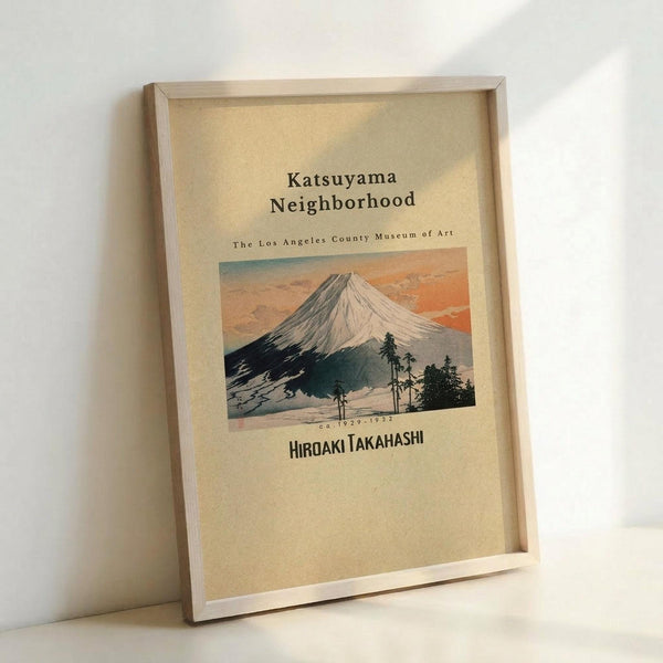 A framed vintage-style poster featuring a Japanese woodblock print of Mount Fuji with snow-capped peak against an orange and blue sky. Dark green trees are silhouetted in the foreground. Text reads "Katsuyama Neighborhood" and "The Los Angeles County Museum of Art" above the mountain image, with "HIROAKI TAKAHASHI" and "ca.1929-1932" below. The poster has a cream-colored background and is displayed in a light wood frame, leaning against a white wall.
