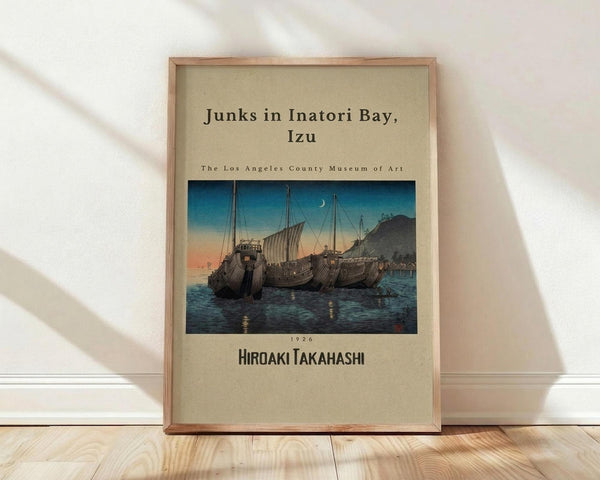 May include: A framed vintage Japanese woodblock print showing traditional sailing junks moored in a bay at twilight. The artwork features deep blue water, orange-pink sunset sky with crescent moon, and dark mountain silhouettes. Text identifies it as depicting Inatori Bay by artist Hiroaki Takahashi from 1926, attributed to Los Angeles County Museum of Art. The print sits in a light wooden frame against white walls on natural wood flooring with soft window light casting diagonal shadows.