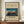 May include: Framed vintage Japanese woodblock print showing traditional sailing junks anchored in a bay at twilight, with blue water, orange sunset sky, and crescent moon. Print features museum exhibition text and artist name. Frame sits on wooden sideboard next to white ceramic vase and books against cream paneled wall.