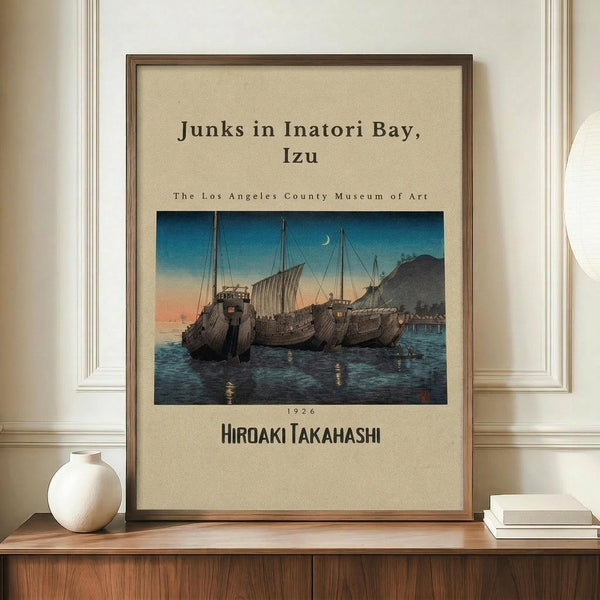 May include: Framed vintage Japanese woodblock print showing traditional sailing junks anchored in a bay at twilight, with blue water, orange sunset sky, and crescent moon. Print features museum exhibition text and artist name. Frame sits on wooden sideboard next to white ceramic vase and books against cream paneled wall.