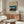 May include: Framed vintage Japanese woodblock print poster showing traditional sailing junks in a bay at sunset, displayed above a mid-century modern walnut credenza with sliding doors. Modern white mushroom table lamp and curved white chair visible in minimalist living room setting with glass oval coffee table and neutral beige walls.