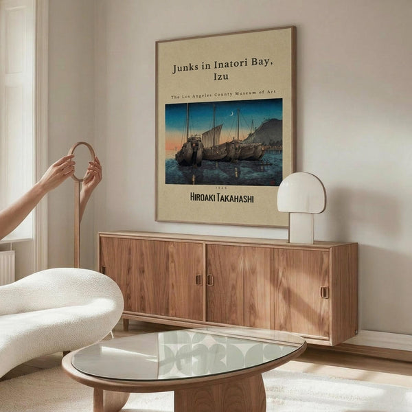 May include: Framed vintage Japanese woodblock print poster showing traditional sailing junks in a bay at sunset, displayed above a mid-century modern walnut credenza with sliding doors. Modern white mushroom table lamp and curved white chair visible in minimalist living room setting with glass oval coffee table and neutral beige walls.