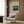May include: Japanese woodblock print poster featuring traditional sailing junks on blue water at sunset, mounted on wood-paneled wall above beige upholstered armchair with geometric patterned pillow. Modern reading corner with brass floor lamp, dark round side table with open book, and natural lighting from window with white curtains.