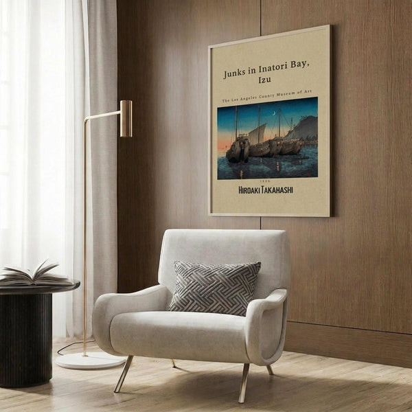May include: Japanese woodblock print poster featuring traditional sailing junks on blue water at sunset, mounted on wood-paneled wall above beige upholstered armchair with geometric patterned pillow. Modern reading corner with brass floor lamp, dark round side table with open book, and natural lighting from window with white curtains.