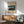 May include: Framed vintage Japanese woodblock print showing traditional sailing junks moored in a bay at twilight, displayed above a mid-century modern wooden credenza with white ceramic lamp. Sage green upholstered chair with wooden arms sits nearby on hardwood floor. Warm sunlight streams across white wall creating geometric shadows. Print features blue-orange sunset sky and dark silhouetted boats on water.
