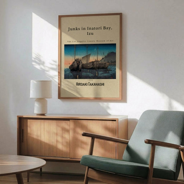 May include: Framed vintage Japanese woodblock print showing traditional sailing junks moored in a bay at twilight, displayed above a mid-century modern wooden credenza with white ceramic lamp. Sage green upholstered chair with wooden arms sits nearby on hardwood floor. Warm sunlight streams across white wall creating geometric shadows. Print features blue-orange sunset sky and dark silhouetted boats on water.