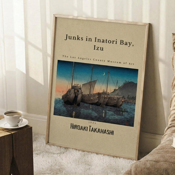 May include: Framed vintage Japanese woodblock print showing traditional sailing junks in a harbor at twilight, with blue and orange sunset sky and crescent moon. The beige matted artwork leans against white wall corner on white textured rug, next to wooden side table with white coffee cup and saucer. Cozy room setting with natural window light and neutral home decor.